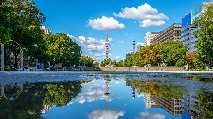 A tower near buildings and trees with its reflection on a body of water