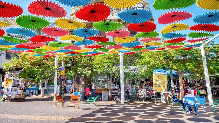 People walking and sitting in front of a building with colorful paper shades above