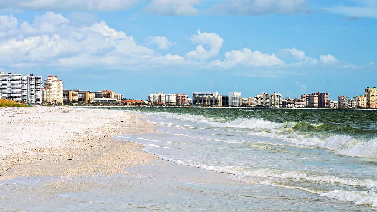 A beach with buildings on the horizon under a blue sky with clouds