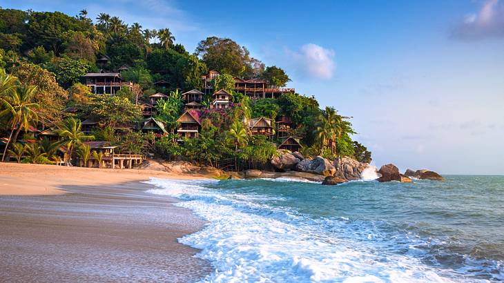 Waves crashing on the shore near a greenery-covered mountain with huts