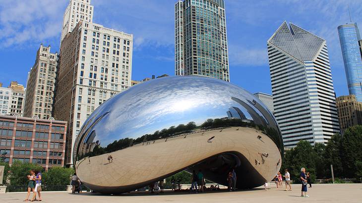 A reflective bean-shaped sculpture with tourists around, against tall buildings