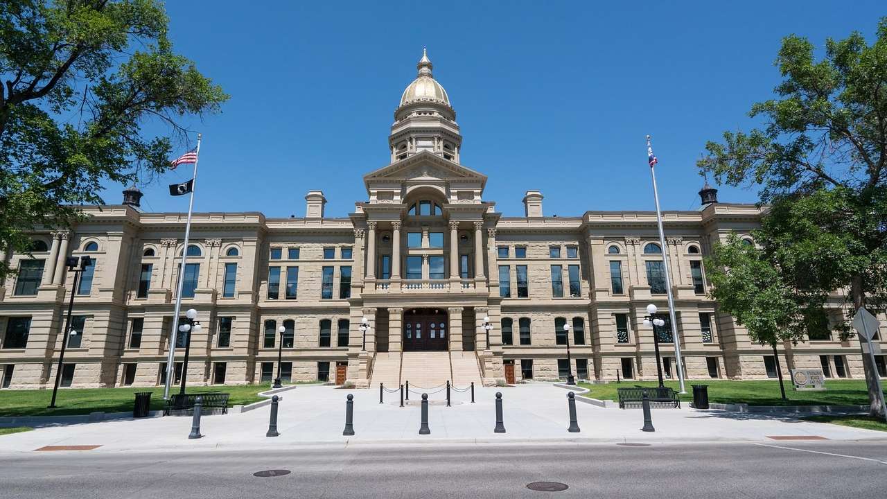 A large state capitol building with a domed roof and steps, flags, and trees in front