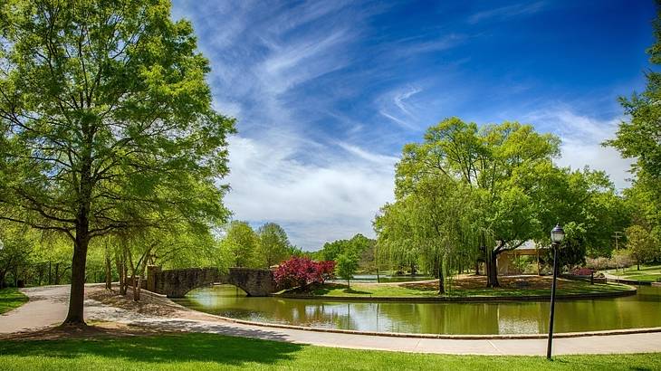 A park with trees, a paved walkway, and a river with a small stone bridge over it