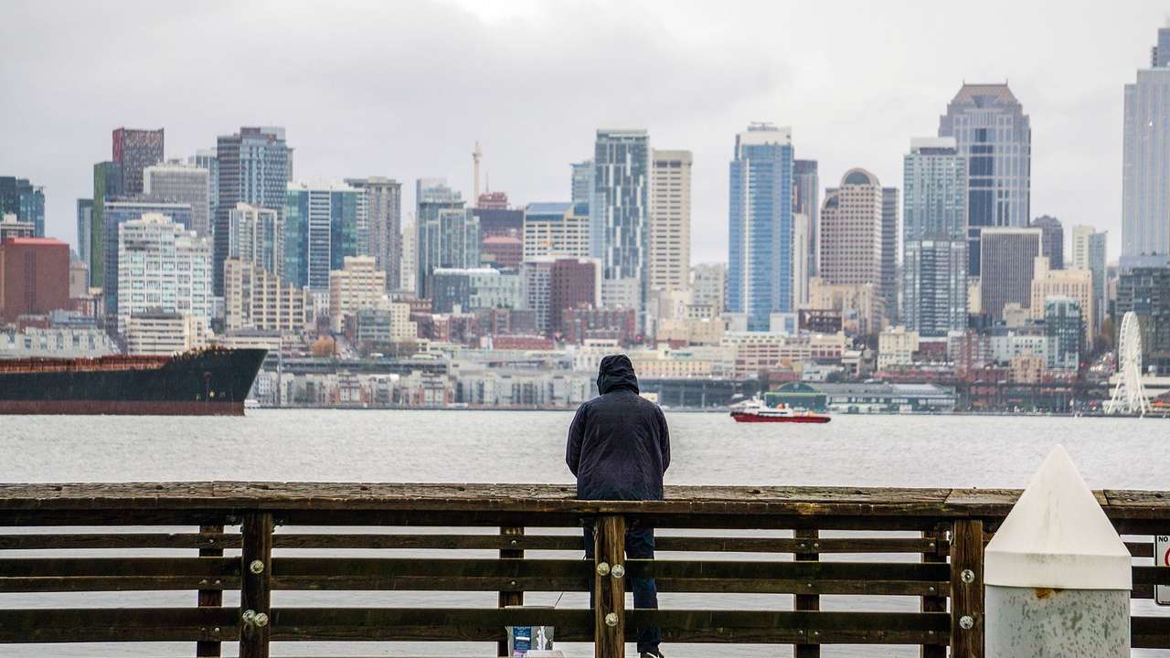 A person in a black raincoat next to the water and city buildings on an overcast day