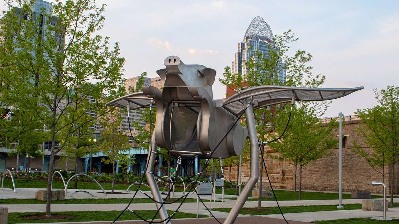 A metal sculpture of a pig with wings in a playground next to trees and buildings