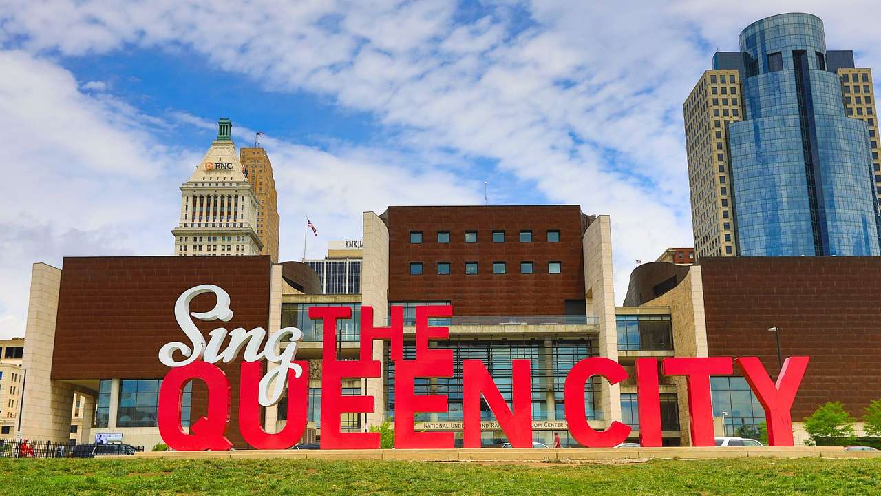 A large red sign that says "Sing the Queen City" on a lawn next to buildings