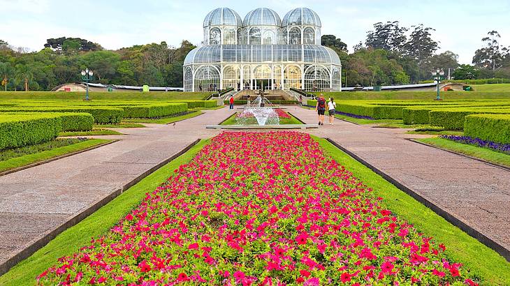 A large flower garden in front of a glass building with three domes and trees behind