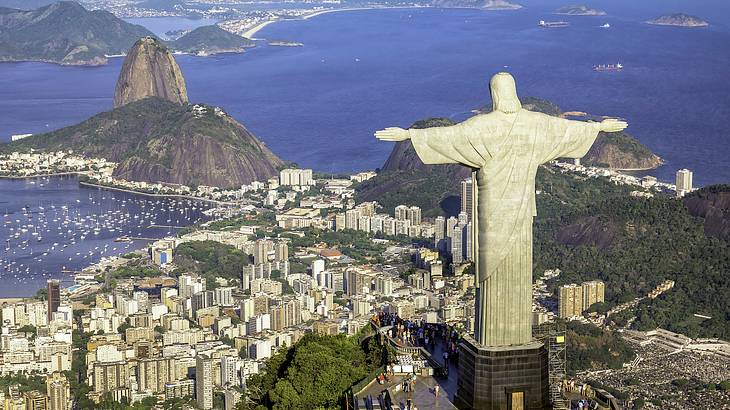 A big statue of Christ on a tall mountain top overlooking buildings and trees below