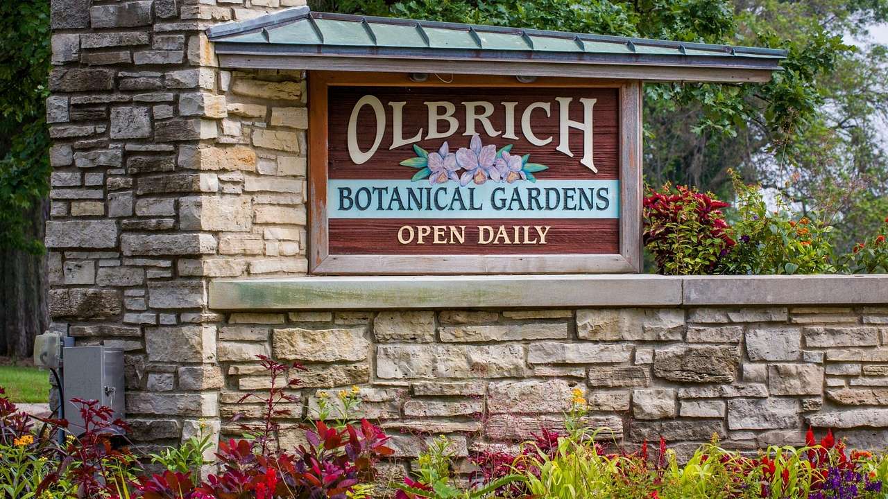 A sign that says "Olbrich Botanical Gardens, open daily" on a stone wall with flowers