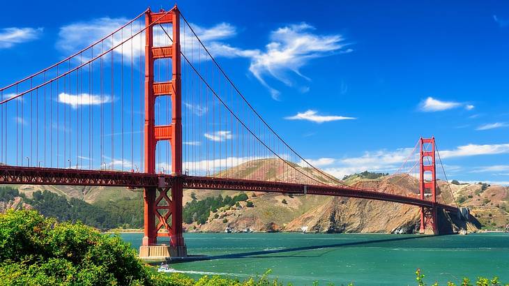 A red suspension bridge with water below it and greenery and mountains surrounding it