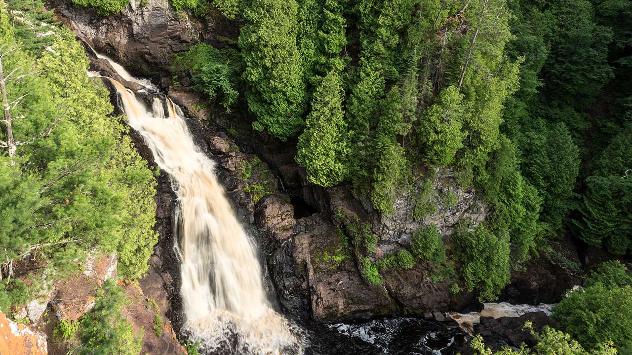 A cascading waterfall between tree-lined cliffs drops into a dark river