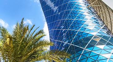 A view of the glass panels of a skyscraper from below with a palm tree on the left