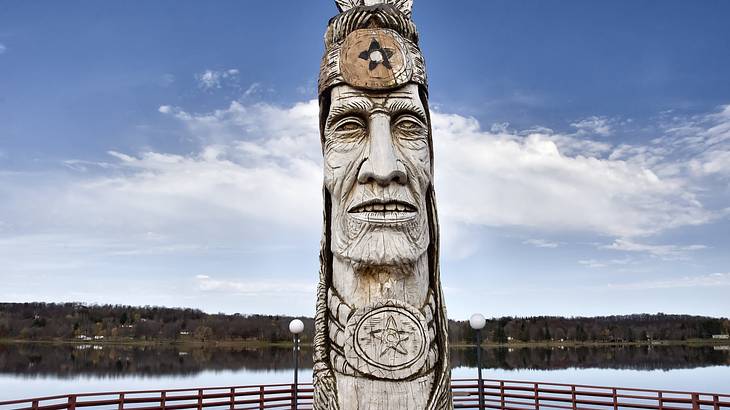 A totem pole featuring the carved face of a Native American placed in front of a lake