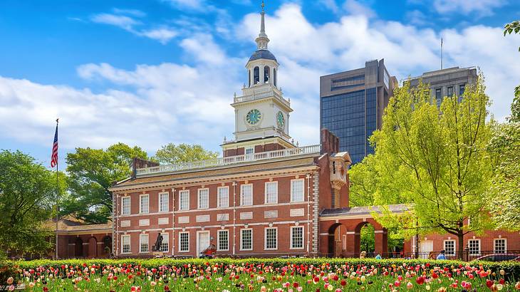 A red brick building with a clock tower next to grass with flowers and a US flag