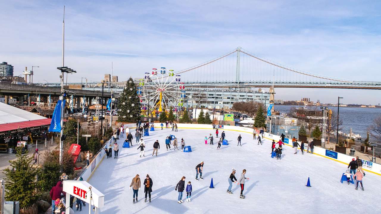 An ice rink with people skating next to a Ferris wheel, bridge, and a river