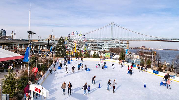 An ice rink with people skating next to a Ferris wheel, bridge, and a river