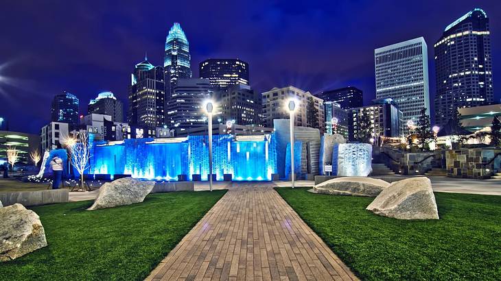 A park with a blue illuminated fountain feature and skyscrapers behind it at night