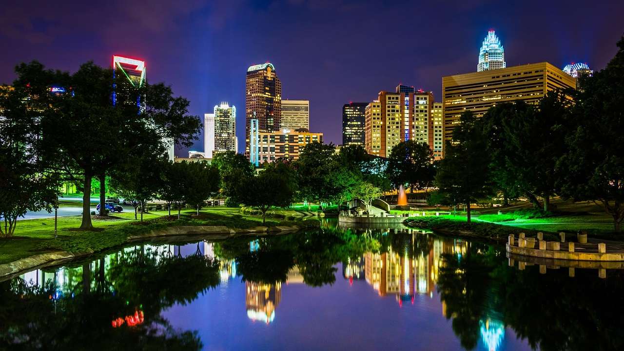 A city skyline illuminated at night with a lake and trees in front of it