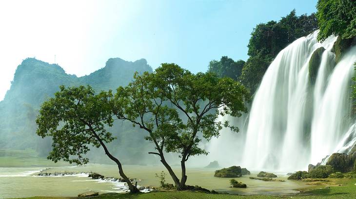 A massive waterfall going off a lush green cliff with mountains and trees around it