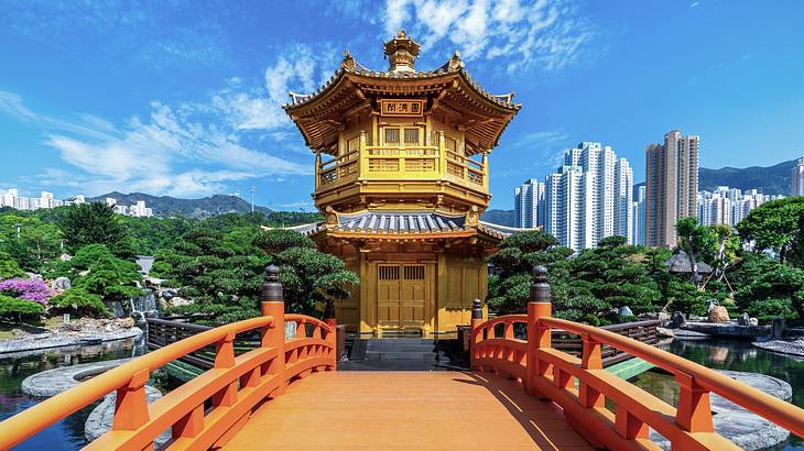 An orange bridge leading to a pagoda near bonsais and skyscrapers in the background