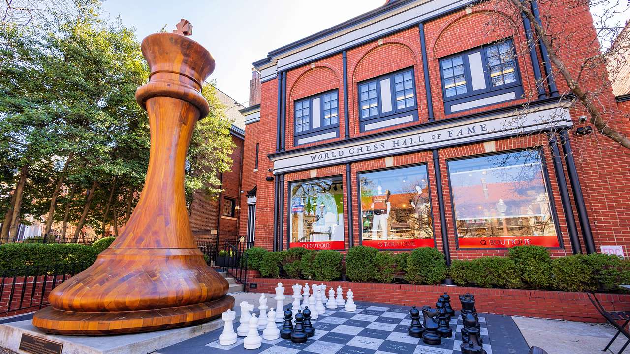 A replica of a tall chess pawn beside a large chess board in front of a building
