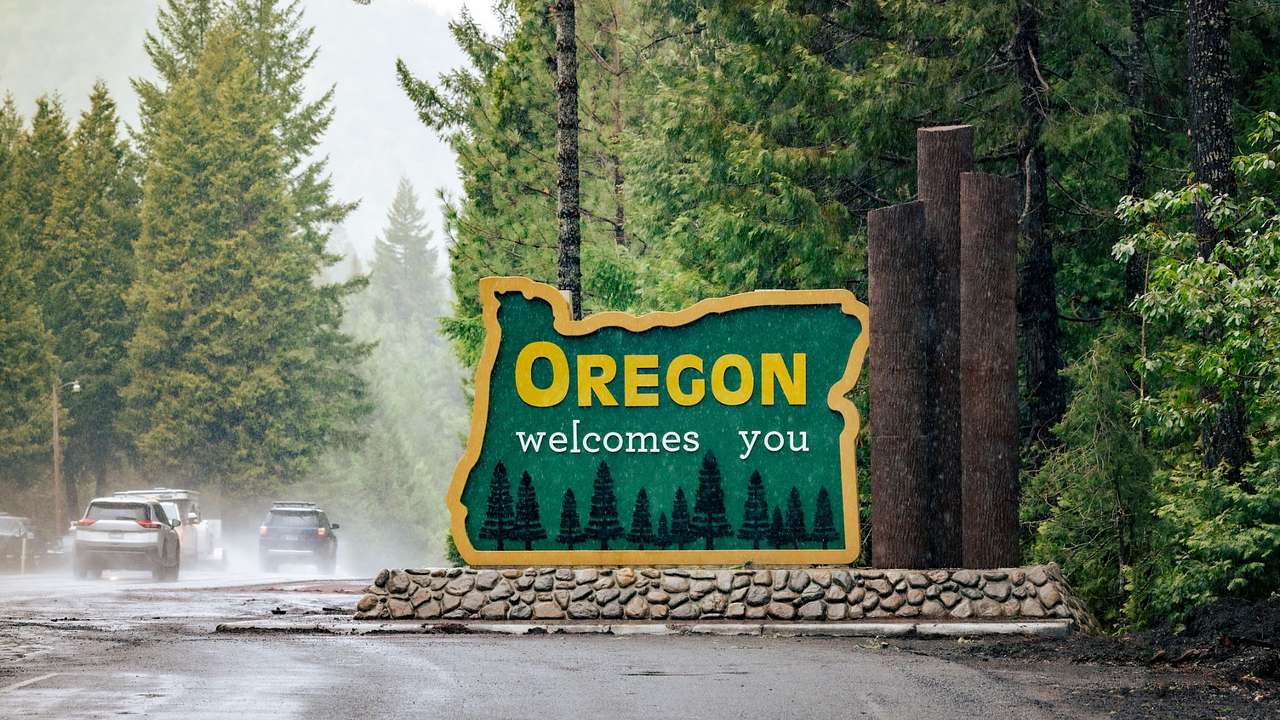 A sign that says "Oregon Welcomes You" next to a road with cars and trees during rain