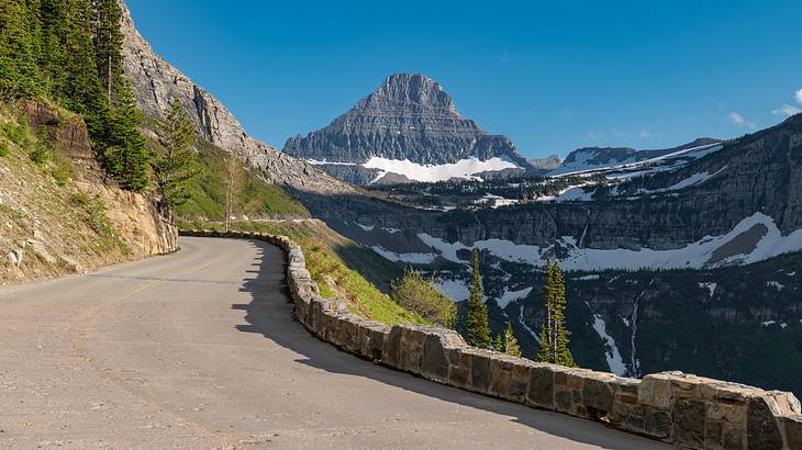A winding mountainside road with a view of a rocky mountain with patches of snow