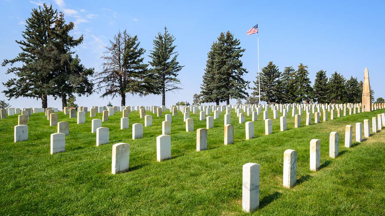 White granite tombstones, pine trees, and a flag of the US on a grassy lawn