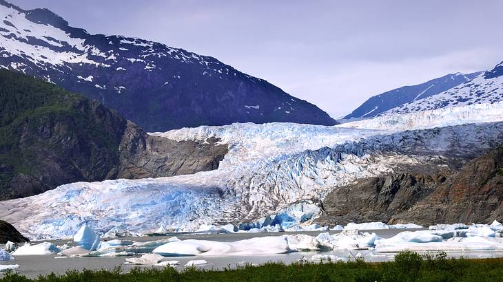 Blue ice formation floating over a hill and into a lake, surrounded by mountains