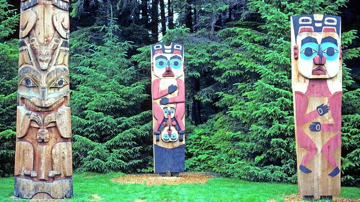 Three multi-colored totem poles with faces carved in them, in front of trees