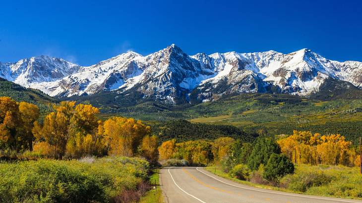 A road through lush greenery next to snow-capped mountains under a blue sky