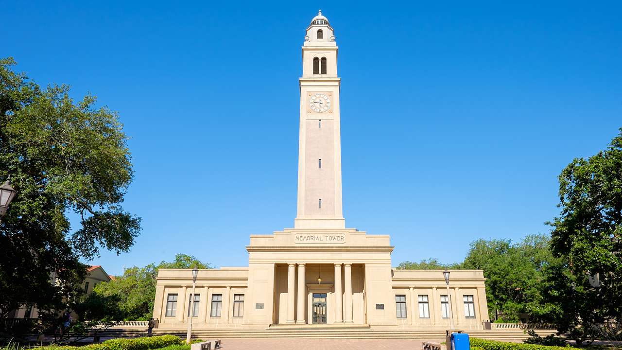 A neoclassical building with a tall tower and trees at its sides, under a clear sky