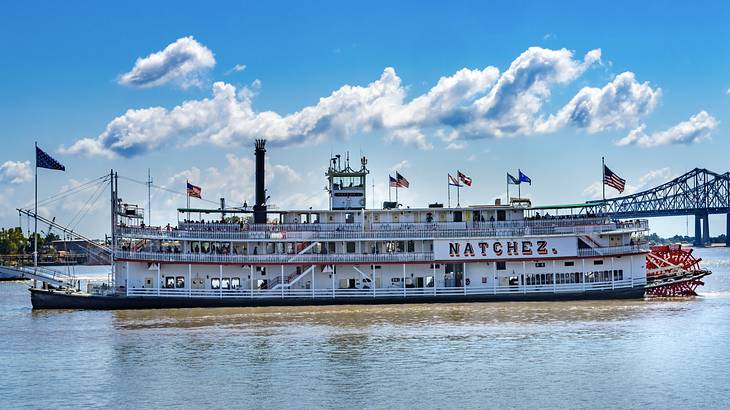 A white steamboat with flags on it on the water with a bridge in the background