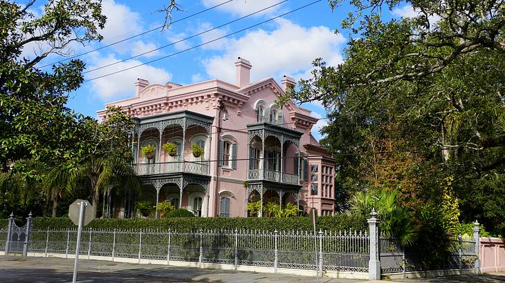 A pink mansion with white iron gate and a road and trees in front