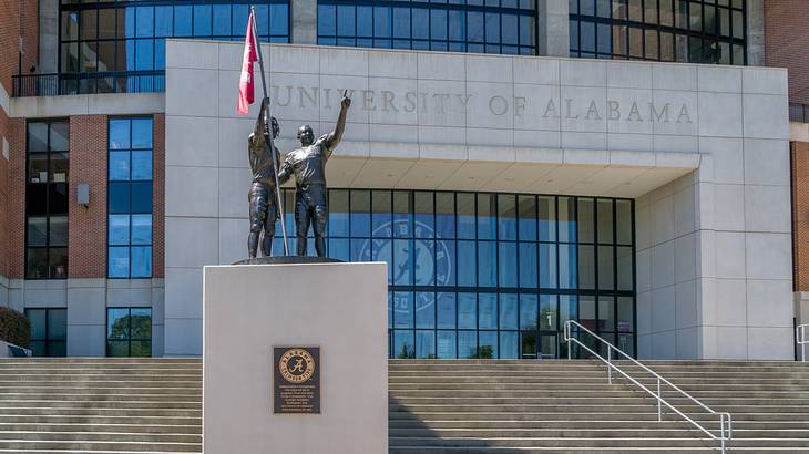 A bronze statue of 2 people holding a flag in front of stairs leading to a building