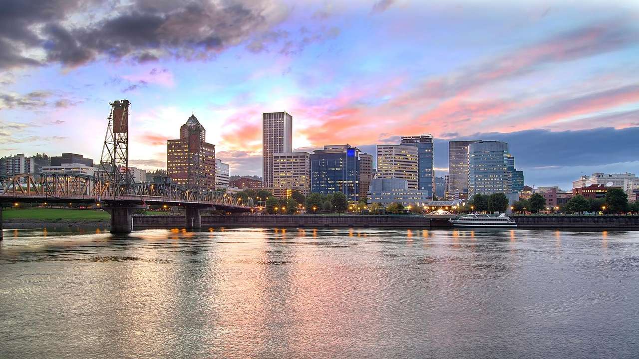 A city skyline illuminated with a river and a bridge in front of it at sunset