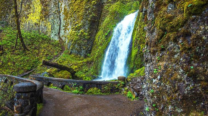 A waterfall flowing over rocks with green moss and a small bridge in front of it