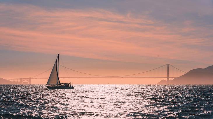 A sailboat on the water with a suspension bridge in the distance at sunset