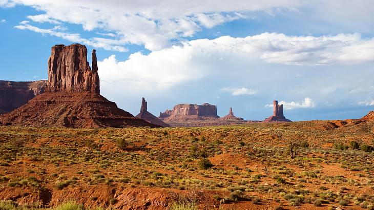 A desert with bushes with a big sandstone hill under a partly cloudy sky