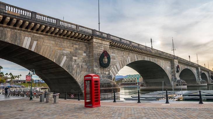 A red telephone booth against an arched bridge under a cloudy sky