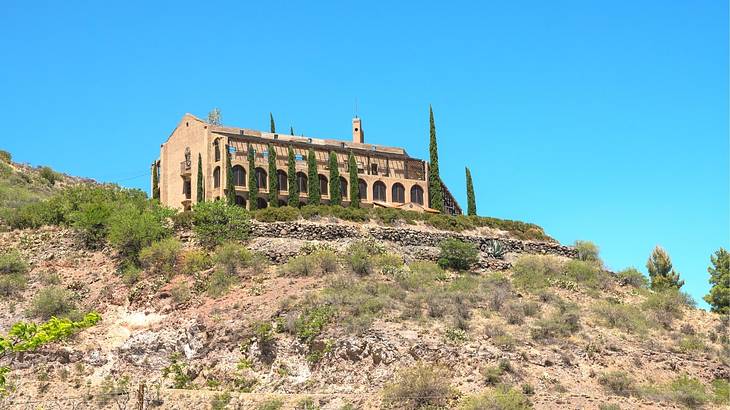 A building with arched windows on top of a rugged hill with bushes against a blue sky
