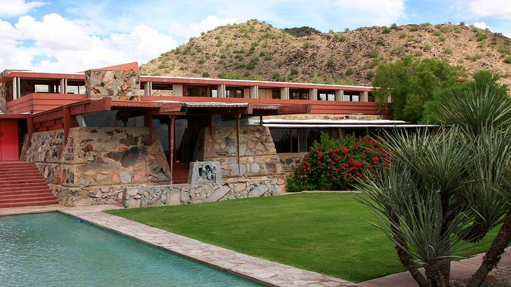 A pool and green grass outside a wide red house against a hill