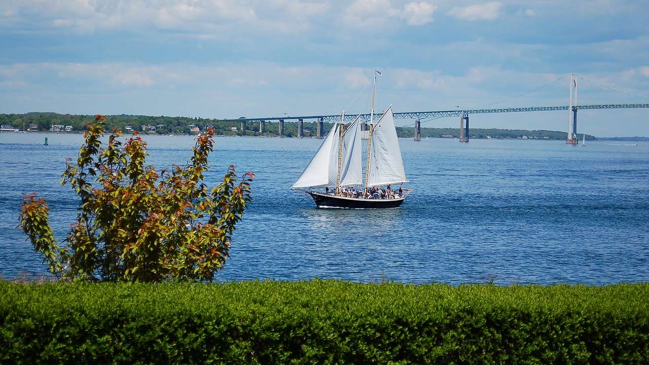 Sailboat on a body of water under a bright, cloudy sky with a bridge in the distance