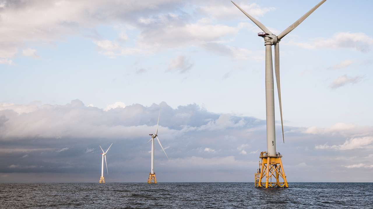 Three large wind turbines rising above the sea against a cloudy, light-blue sky