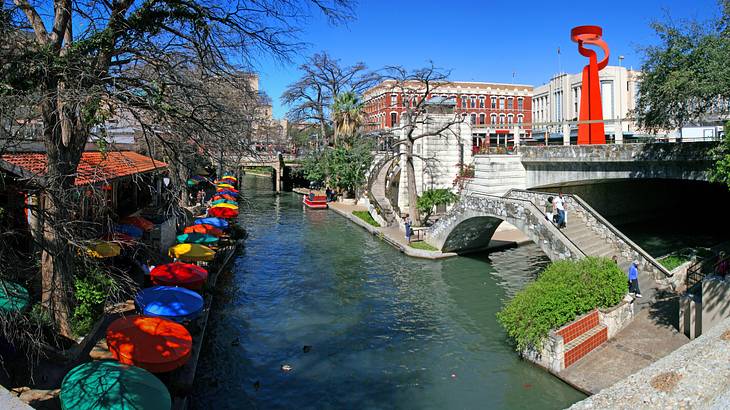 A blue river with colorful patio umbrellas on the left, walkway bridge on the right