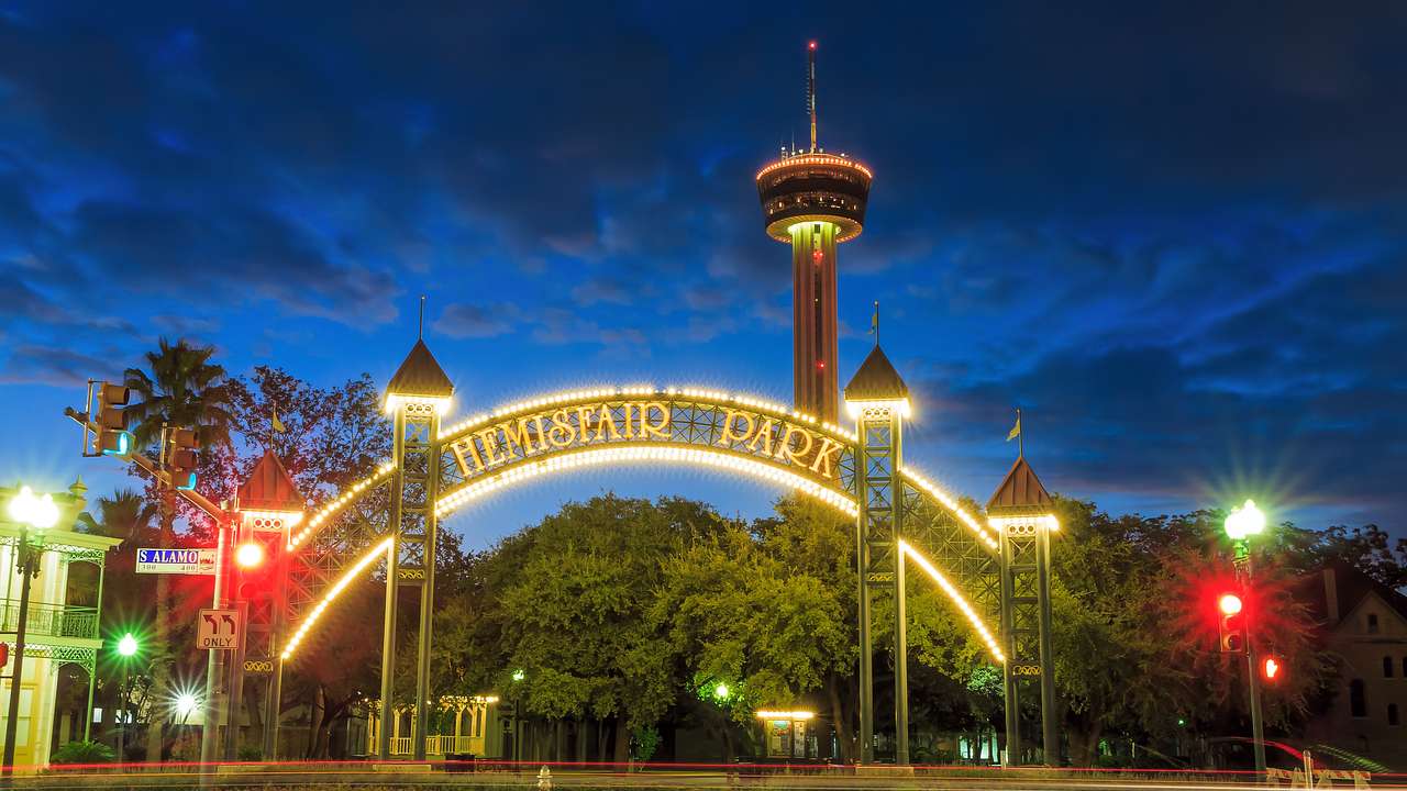 Wide round illuminated gate saying "Hemisfair Park" in front of a sky tower at night