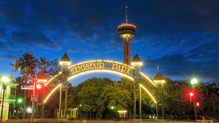 Wide round illuminated gate saying "Hemisfair Park" in front of a sky tower at night