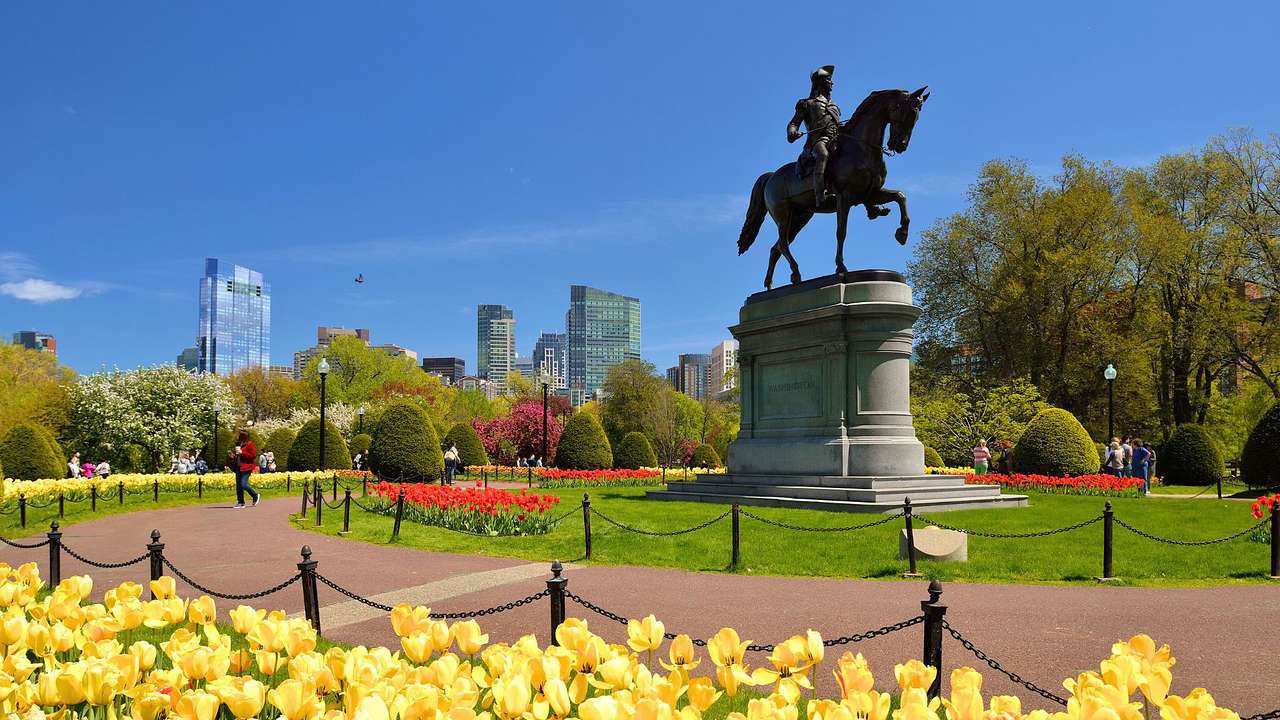 A park with flowers, a walking path, trees, and a statue of a man on a horse