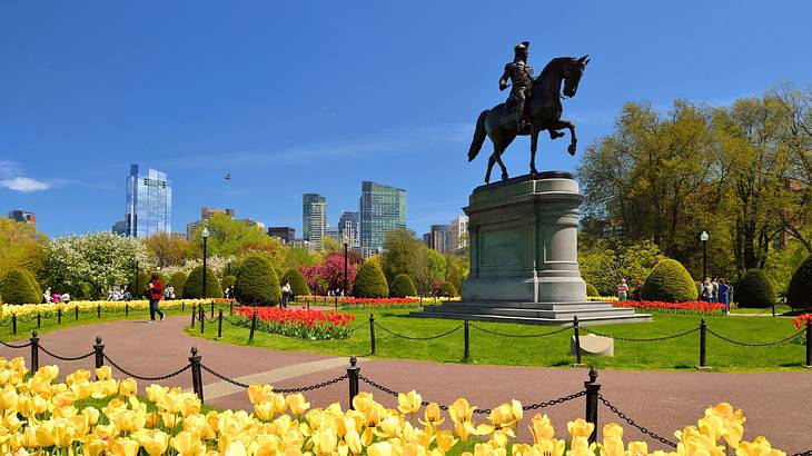 A park with flowers, a walking path, trees, and a statue of a man on a horse