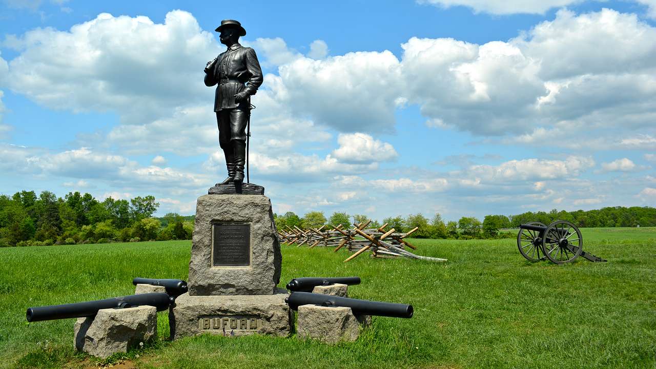 A bronze statue on a stone pedestal with four canons at its base in a grassy field
